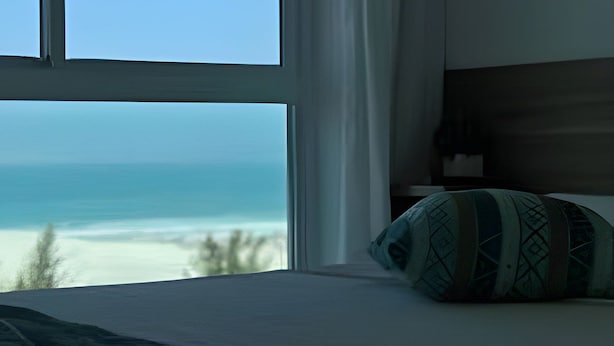 A bedroom with a large window, a bed with a patterned pillow, and a view of the ocean. - Flat Mandai Cabo Frio (Cabo Frio)