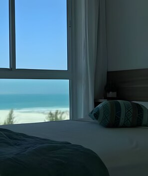 A bedroom with a large window, a bed with a patterned pillow, and a view of the ocean. - Flat Mandai Cabo Frio (Cabo Frio)