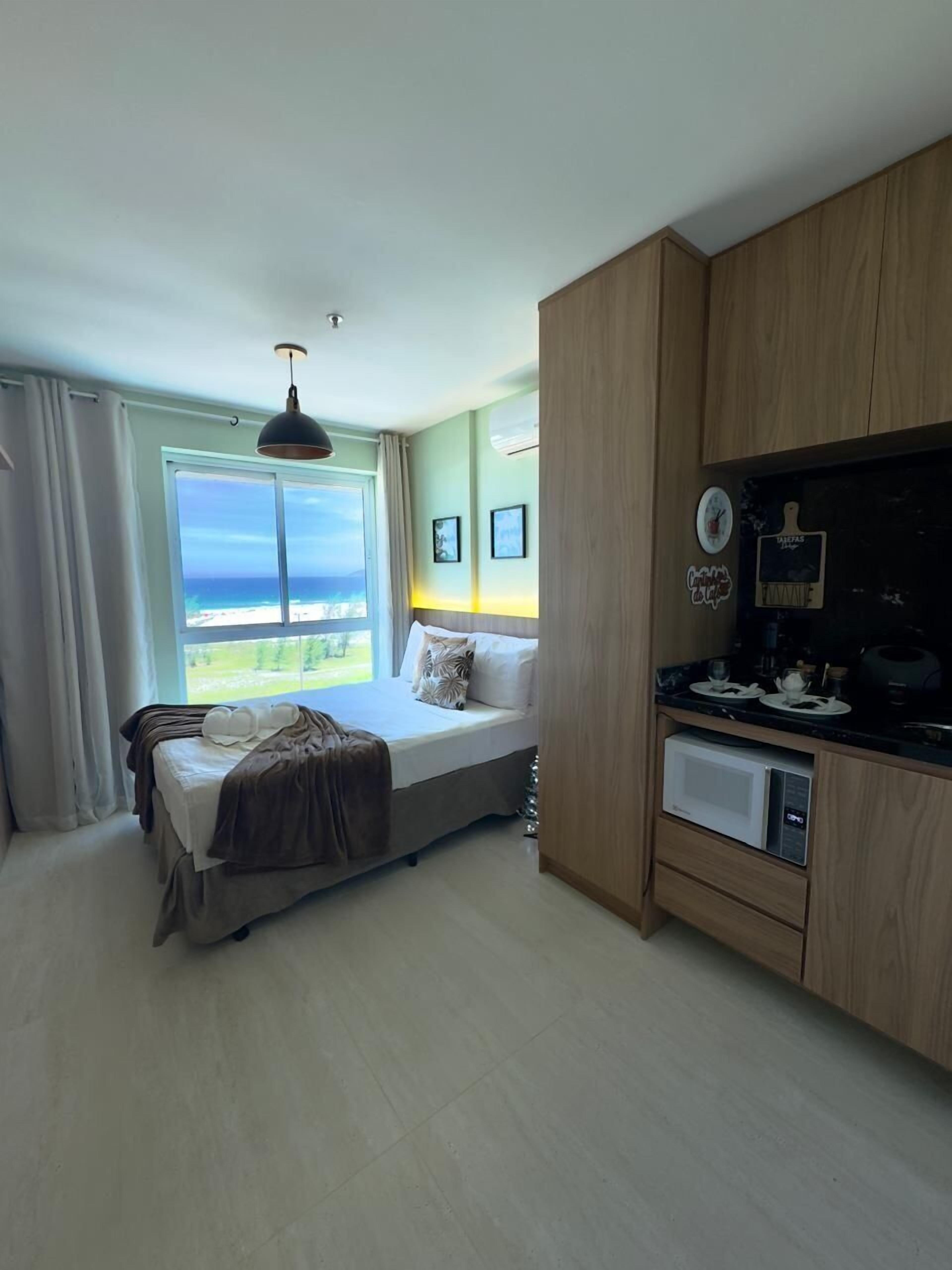 A bedroom with a bed, a view of the ocean, a kitchenette, and a ceiling light.