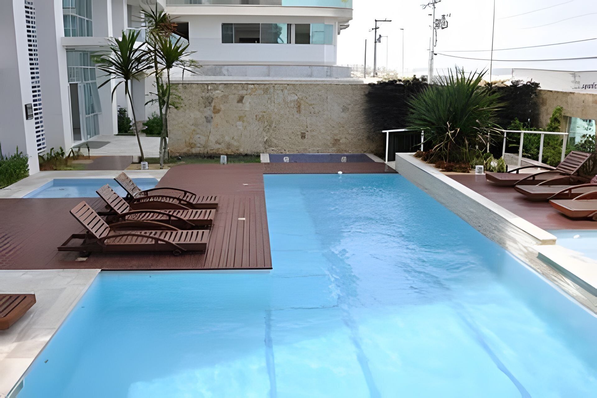 Una piscina amb gandules, una paret de pedra i edificis moderns al fons.