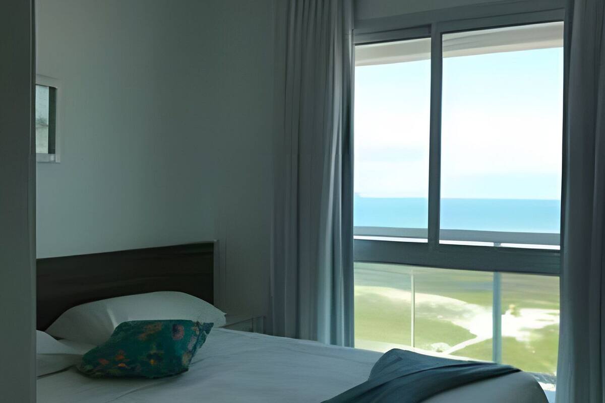 A hotel room with a bed, a view of the sea, and a window with curtains.