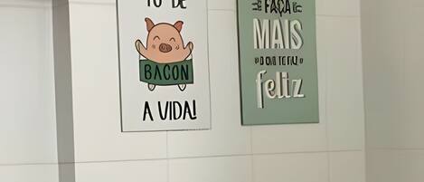 Two wall-mounted signs in a hallway, one with a cartoon pig and the text 'Tô de bacon a vida!' and another with the text 'Faca mais feliz.'