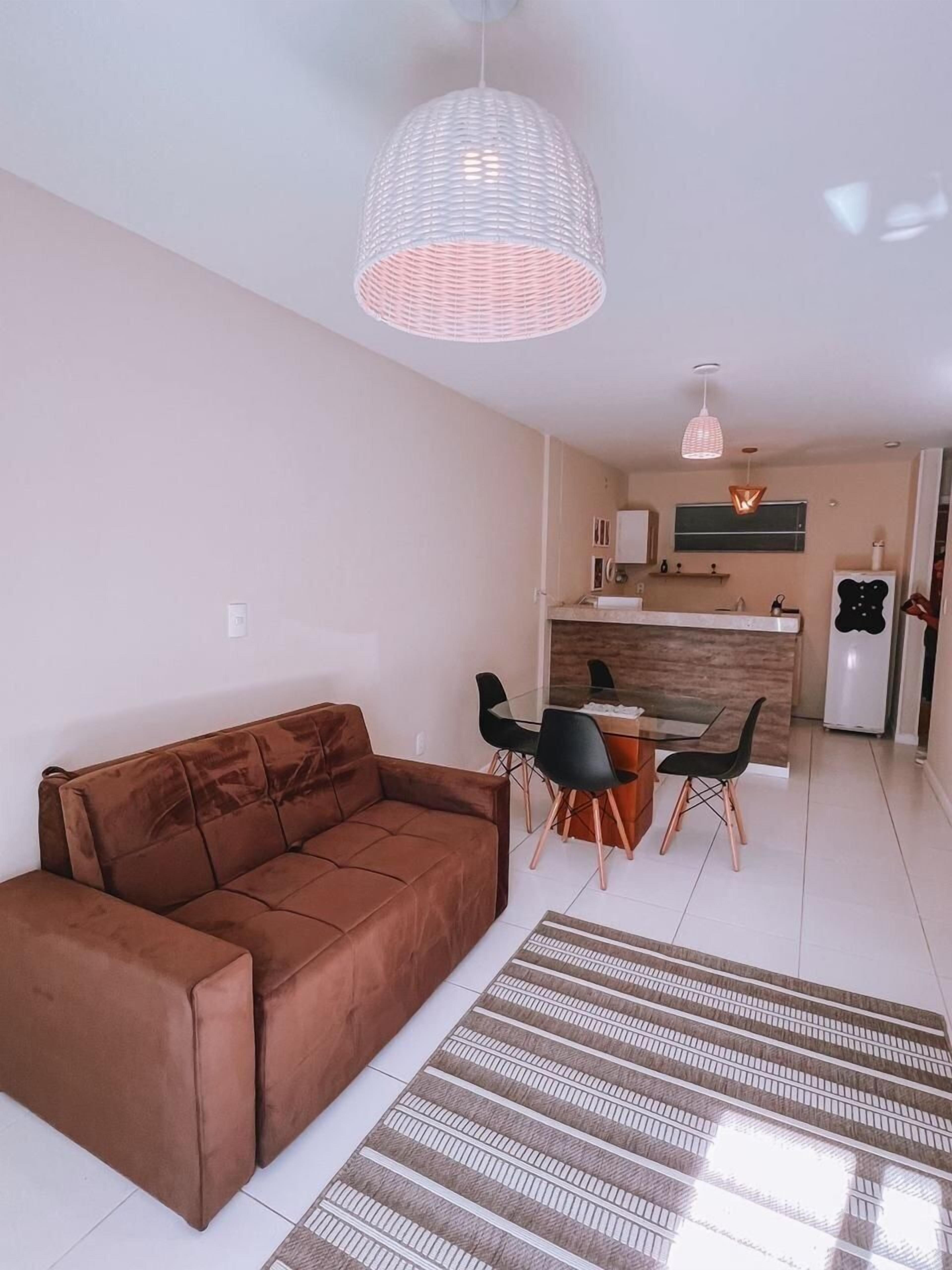 A modern living room with a brown sofa, a glass dining table with black chairs, a kitchen in the background, and a patterned rug.