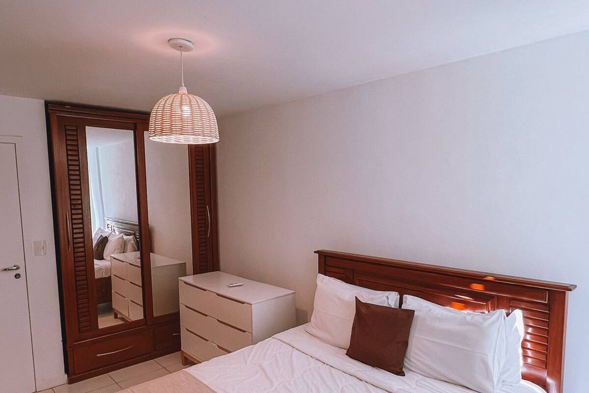 A bedroom with a wooden bed, a bedside table, a dresser, a mirror, and a hanging light fixture.