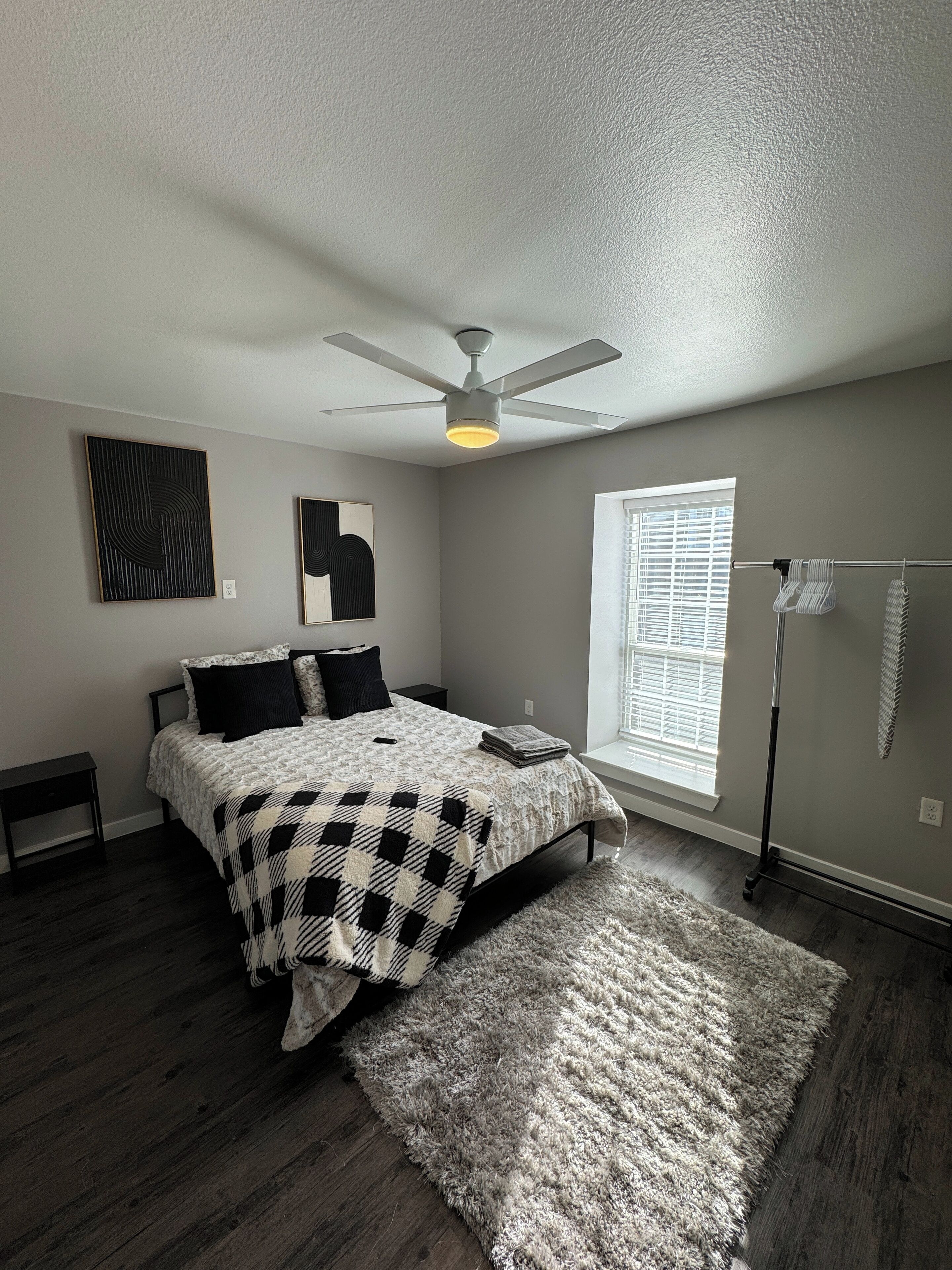 A bedroom with a bed, a ceiling fan, artwork on the wall, a window with blinds, and a floor lamp.
