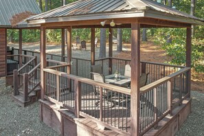 Un belvédère en bois avec une table et des chaises, entouré d’une zone recouverte de gravier et d’arbres.