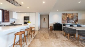 Una cocina moderna con armarios blancos, una isla central con taburetes, un área de comedor con un cuadro grande y una puerta visible que conduce a otra habitación.