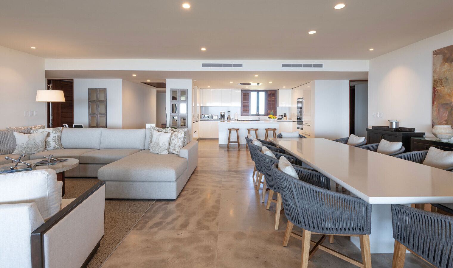 Una sala de estar moderna con un sofá modular, un comedor con una mesa larga y sillas, y una cocina al fondo.