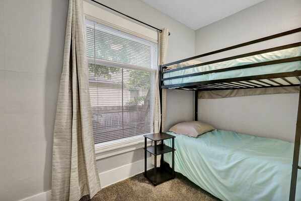 A bunk bed with a small shelf, a window with blinds, and a carpeted floor.