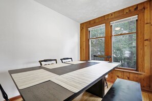 A dining area with a long table, three chairs, and a bench. The room has wooden walls and large windows with a view of trees. - Central | Steps to MSU | Quick Hwy Access | Quiet (Lansing)
