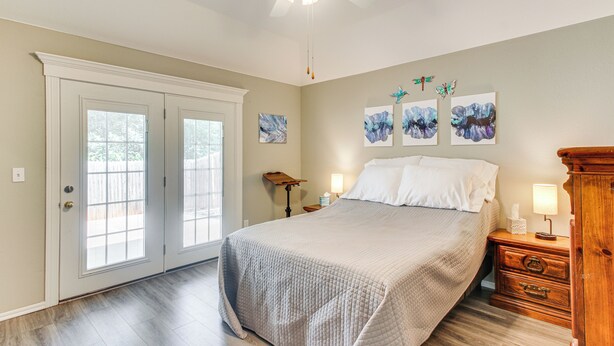 Une chambre à coucher comprenant un lit, une table de chevet, une commode en bois, un ventilateur de plafond et des œuvres d’art accrochées au mur.