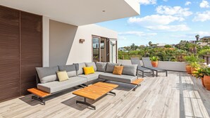 Uma área externa moderna com um sofá modular cinza, mesas de madeira e plantas em vasos.