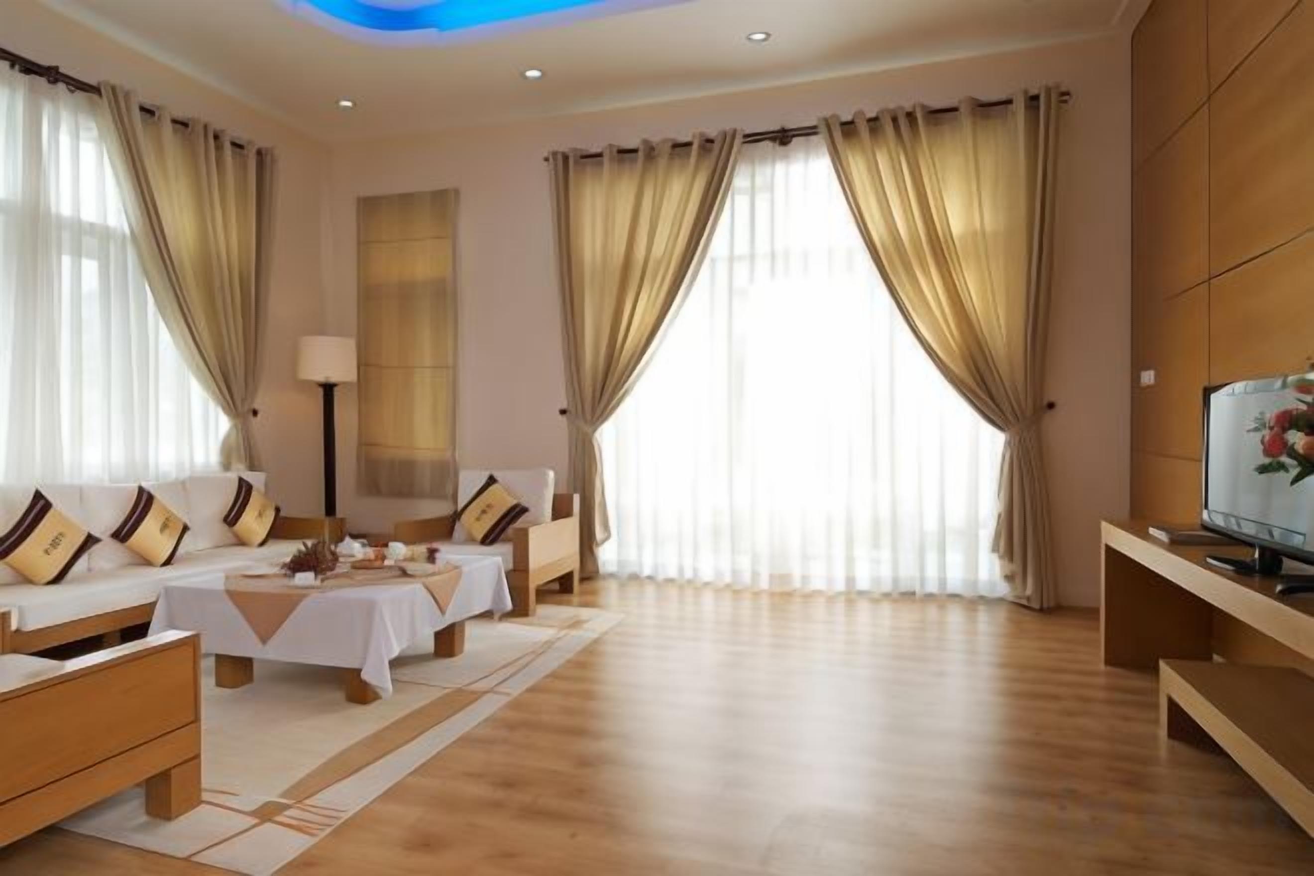 A hotel room with a sofa set, a table, a TV, and large windows with curtains.