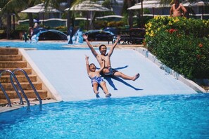 Una piscina con tobogán de agua, escalones para entrar y una palmera al fondo.