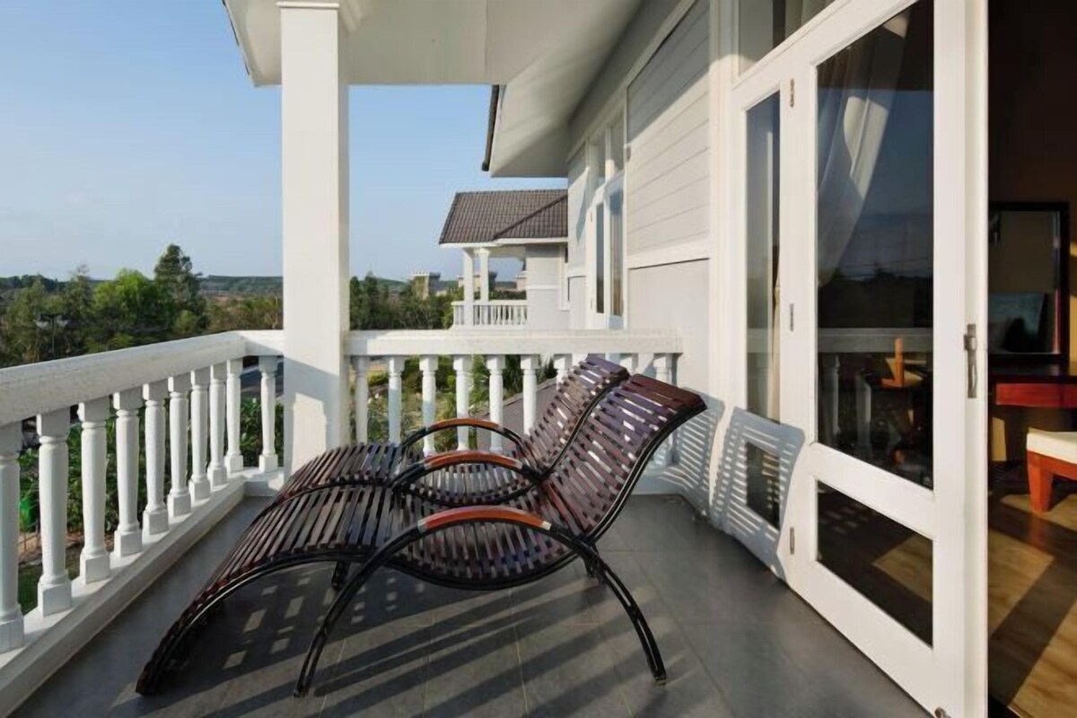 Een balkon met twee stoelen en een tafel, witte leuningen en uitzicht op een huis en bomen.