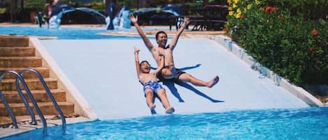 A swimming pool with a water slide, steps leading into the pool, and umbrellas in the background.