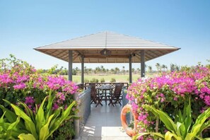 Een overdekt terras met een tafel en stoelen, omringd door weelderige begroeiing en felroze bloemen.