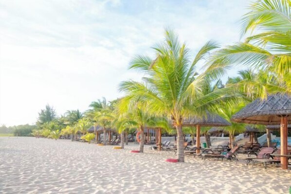 A beach with rows of palm trees and thatched-roof huts, each with a row of sun loungers. - Sea Links Beach Villa (Phan Thiet)