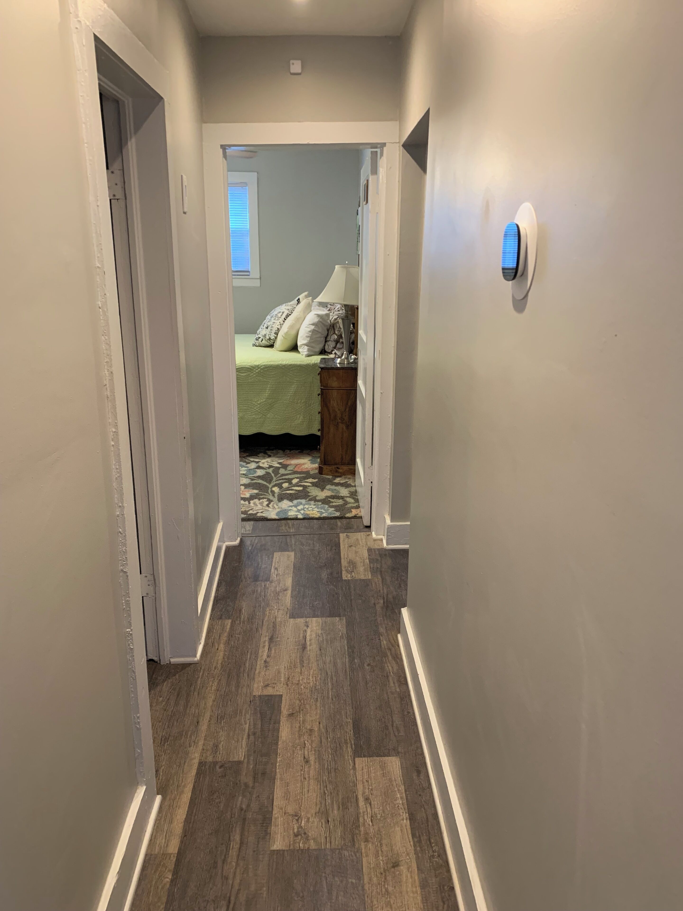 A hallway with a wooden floor leading to a bedroom with a green bedspread and a nightstand.