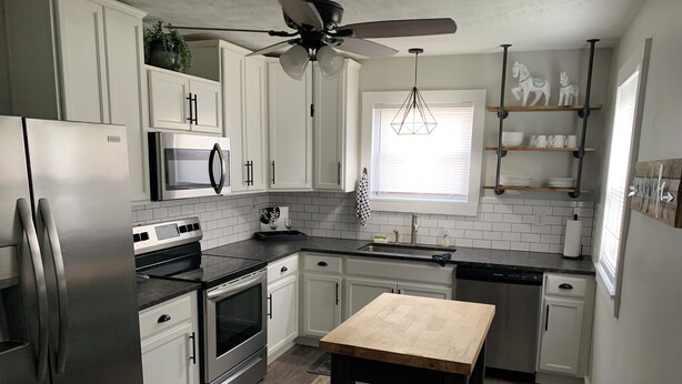 Una cocina moderna con armarios blancos, electrodomésticos de acero inoxidable, una mesa de madera y un ventilador de techo.