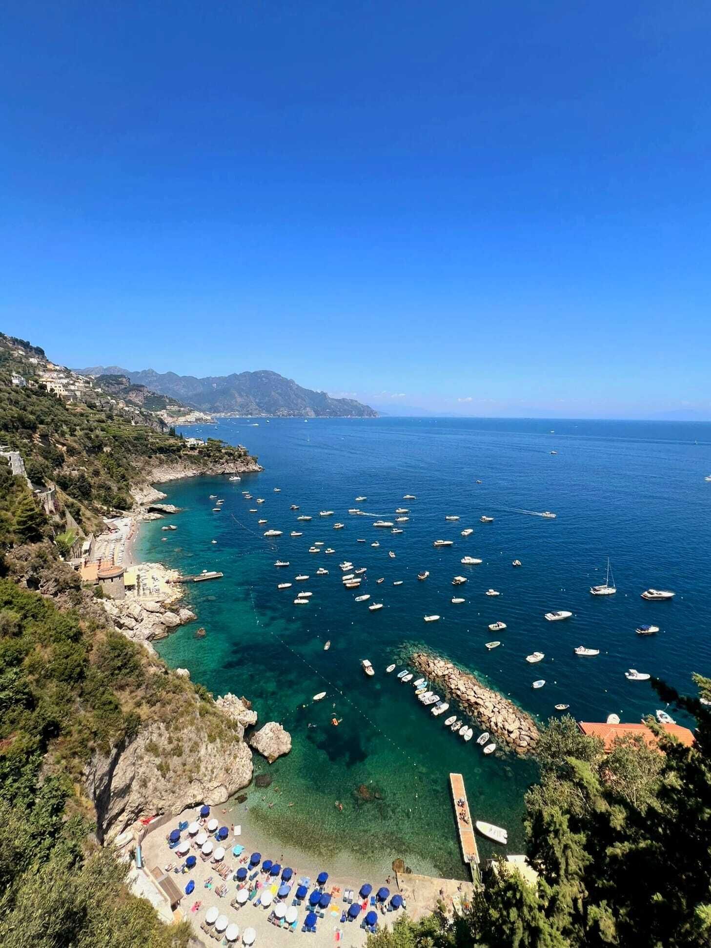A coastal area with clear blue waters, boats, and a beach with umbrellas.