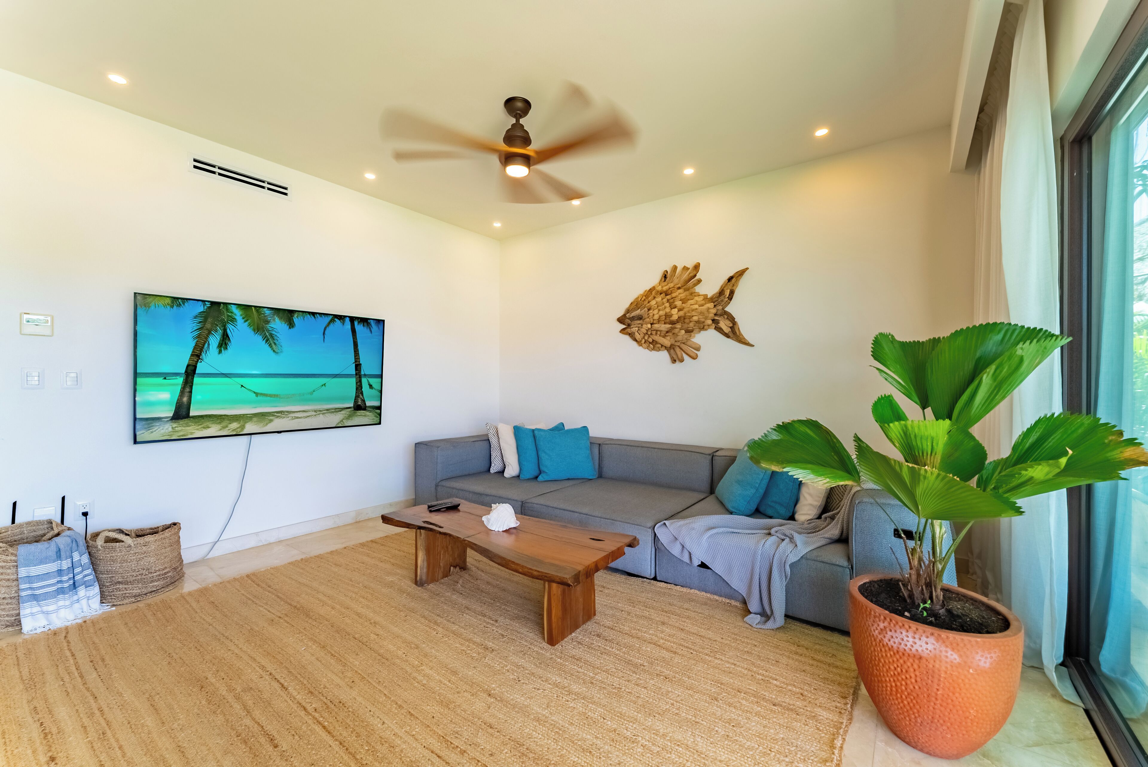 A modern living room with a wooden coffee table, a grey sofa with blue cushions, a large flat-screen TV displaying a beach scene, a ceiling fan, and a large potted plant.