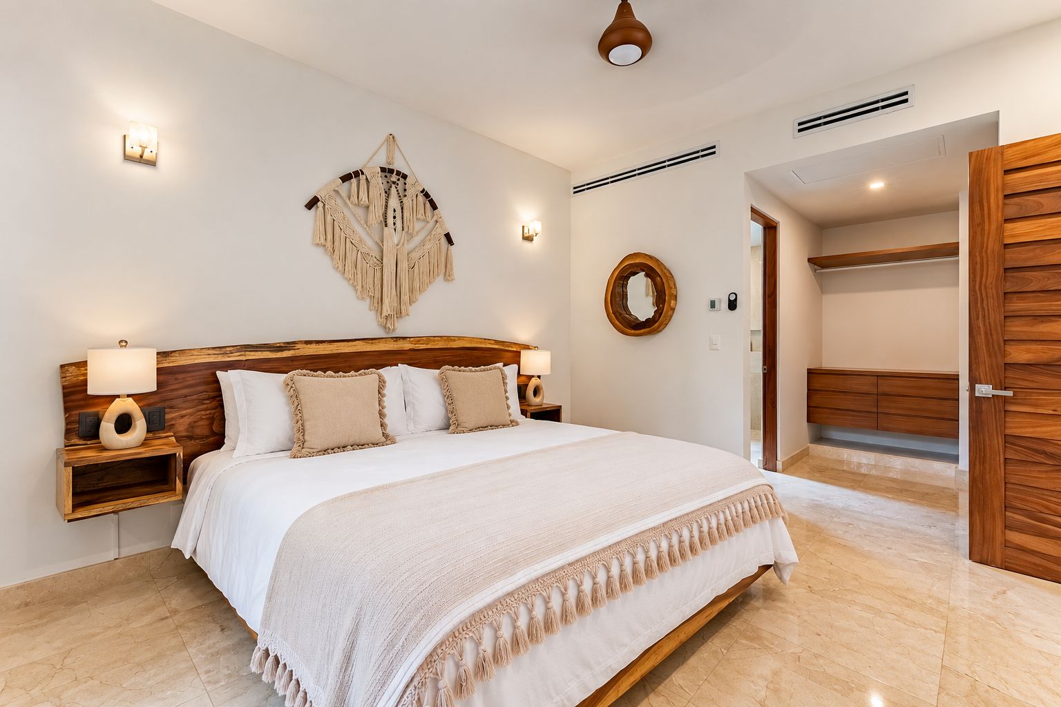 A bedroom with a wooden headboard, a large bed with white bedding, two bedside tables with lamps, a macrame wall hanging, a round mirror, and a wooden wardrobe.