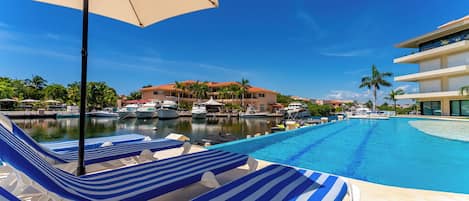 A pool area with lounge chairs and an umbrella, a marina with boats, and a building with a red roof.