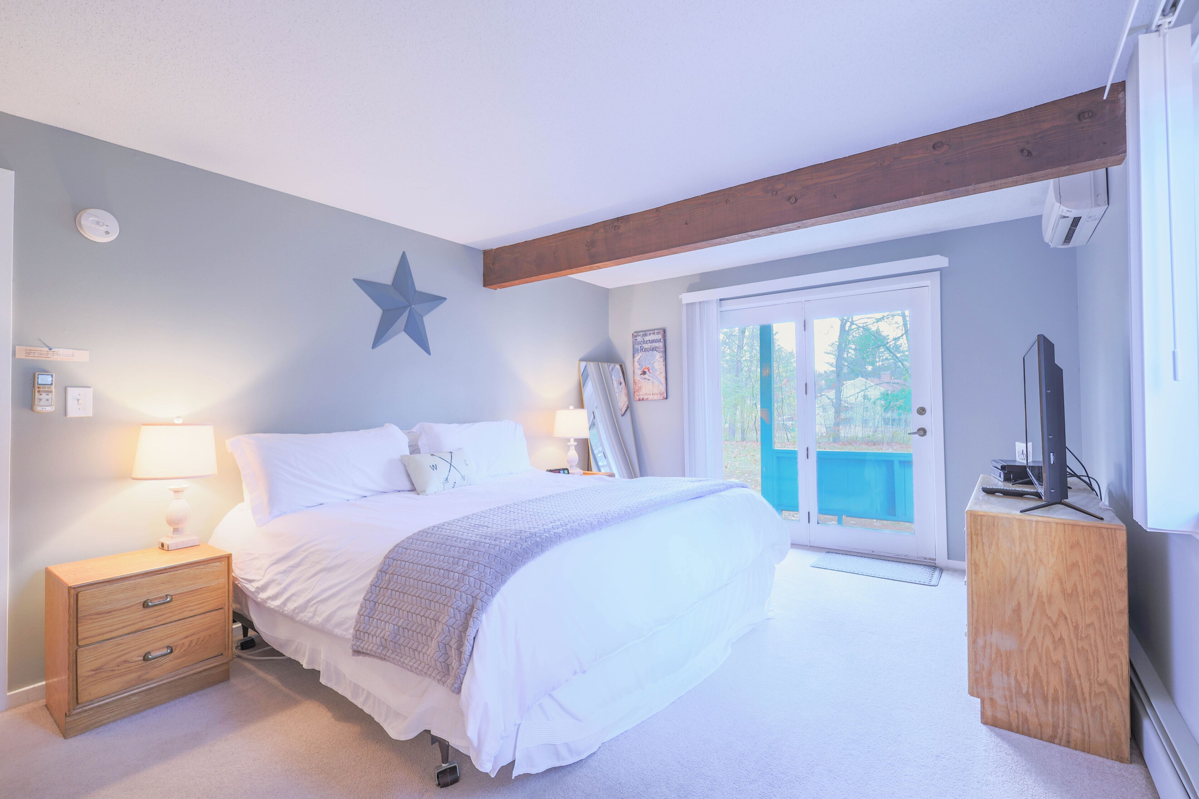 A bedroom with a bed, bedside lamps, a star decoration, a wooden headboard, a television, and a sliding glass door to the outside.