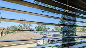 A view through window blinds of a residential area with a brick house, a lawn, and a body of water in the background. - Perfect Family Escape: Playground, Fire Pit, Games & Boat Parking Near Kemah! (Bacliff)