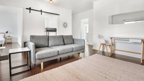 A modern living room with a grey sofa, a small side table, a white desk, and a large window. - Bonbeach Beauty: Relaxation Haven in Bonbeach! (Bonbeach)