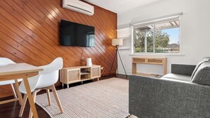 A modern living room with a wooden wall, a flat-screen TV, a sofa, a dining table with chairs, and a lamp. - Bonbeach Beauty: Relaxation Haven in Bonbeach! (Bonbeach)