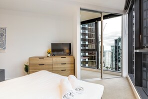 Une chambre d’hôtel moderne avec un lit, un téléviseur posé sur une commode en bois, une grande porte-fenêtre coulissante donnant sur un balcon et une vue sur des immeubles de grande hauteur.
