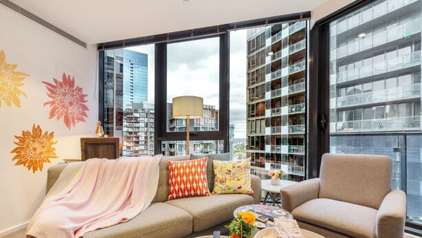 Un salón moderno con sofá, sillón y mesa de centro, ventanas amplias con vistas a la ciudad y una estantería.