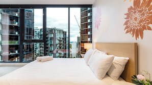 Una habitación de hotel moderna con una cama grande, una mesita de noche con lámpara y vistas a edificios circundantes.
