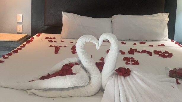 Una cama impecablemente hecha con una toalla dispuesta en forma de corazón y pétalos de rosa.