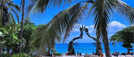 A public square with palm trees and a sculpture, benches, and a view of the sea.
