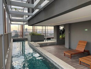 A modern indoor pool area with a wooden lounge chair and a small table, surrounded by concrete walls and a glass railing. - On Melo Alves R (São Paulo)