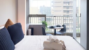 Et hotelværelse med en seng, et lille bord med en stol, en balkon med udsigt til bygninger, og et vindue med persienner.