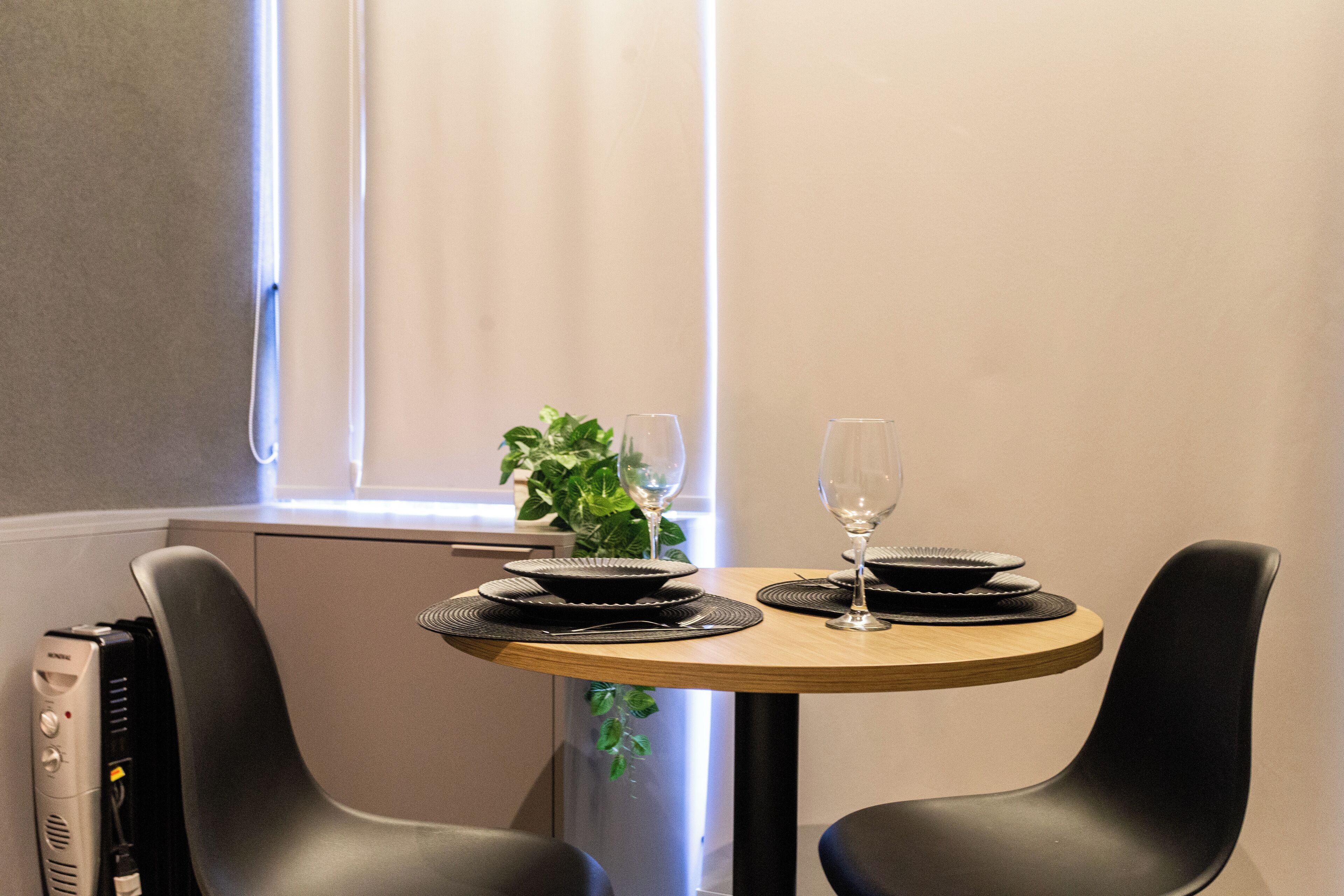 A dining table with black plates and a wine glass, surrounded by black chairs, in a room with a window and a plant.