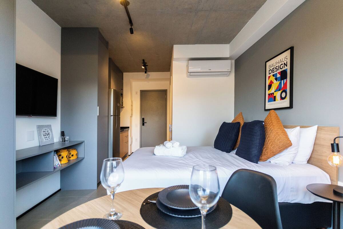 A hotel room with a bed, a TV, a dining area with a table and chairs, and a wall-mounted air conditioner.
