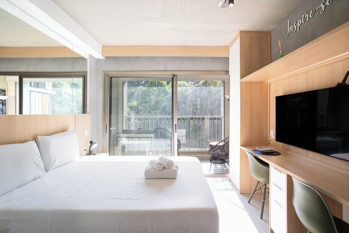 A modern hotel room with a large bed, a desk with a flat-screen TV, and a balcony with a view of trees.