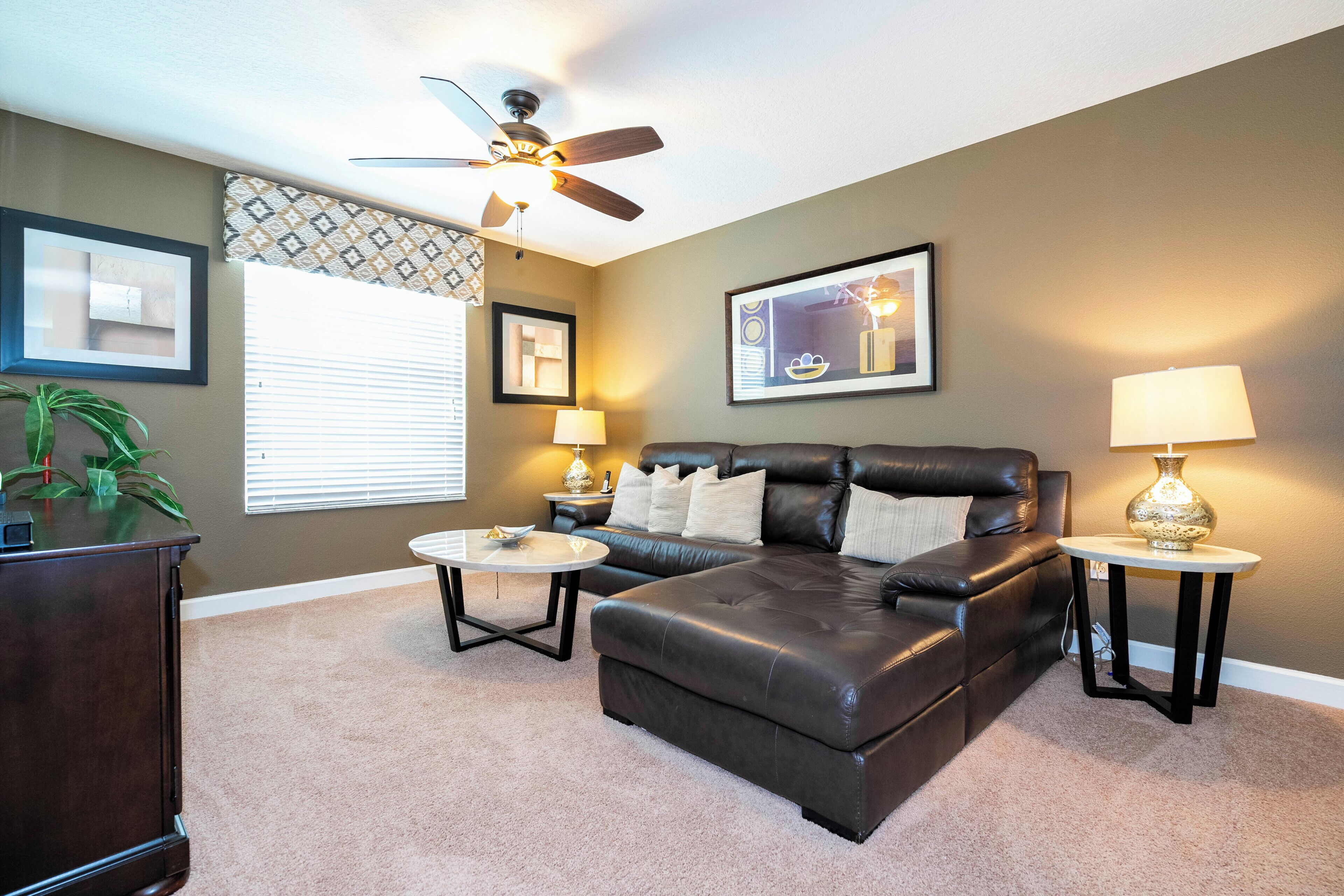 A living room with a leather sofa, two side tables with lamps, a ceiling fan, and framed artwork on the walls.
