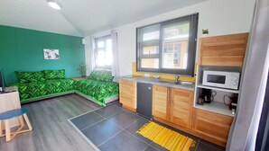 A compact living space with a kitchenette, a bed with green patterned bedding, a small dining area, and a window with blinds.