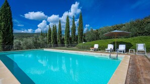 Uma piscina retangular com água azul cristalina, rodeada por um deck ladrilhado. Há três cadeiras de sol brancas com guarda-sóis, e uma cerca de metal delimitando a área. Árvores verdes altas aparecem ao fundo.