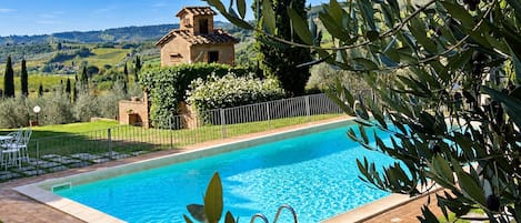 Una piscina con vista a un paisaje campestre, una casa pequeña y olivos.