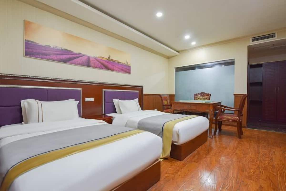 A hotel room with two beds, a wooden floor, a desk with a chair, and a large window.