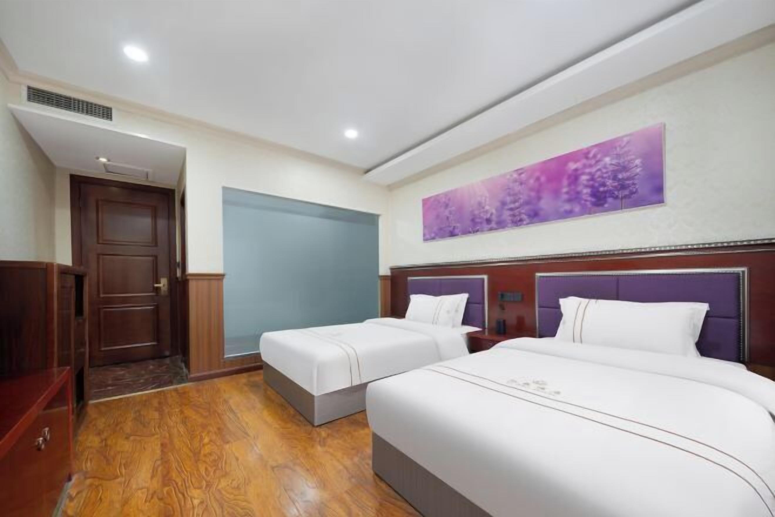 A hotel room with two beds, a wooden floor, a door, and a wall with a purple artwork.