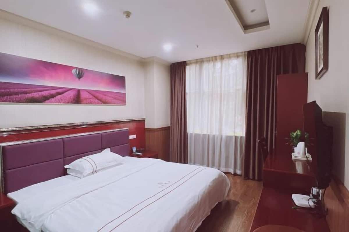 A hotel room with a bed, a painting, a desk, a chair, a TV, and a window with curtains.