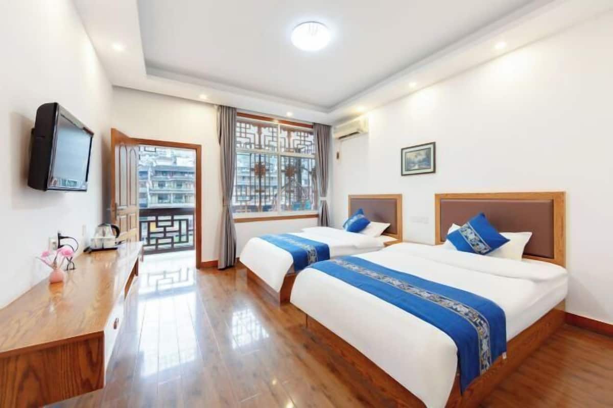 A hotel room with two beds, a wooden desk, a television, and a window with a view of buildings.